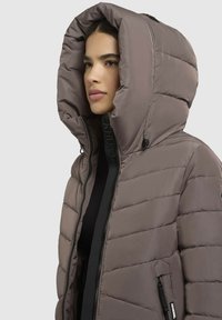 Veste beige rembourrée avec une grande capuche, dotée d'une fermeture éclair à l'avant et d'une poche latérale. La texture matelassée apporte isolation et chaleur.