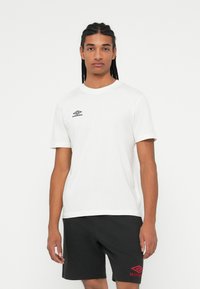 Mladenič s pletenicami, oblečen v belo majico Umbro in črne kratke hlače Umbro, na enobarvni beli ozadju.