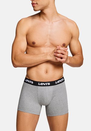 Oberkörperfreier Mann mit muskulösem Körperbau, trägt graue Levi’s Boxershorts, verschränkt die Hände vor der Taille und schaut zur Seite.