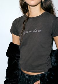 Črna kratka majica s kratkimi rokavi z napisom "Made On" v bleščečih okrasnih elementih. Nosena s črno bunda iz umetnega krzna.