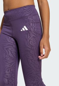 Leggings violets taille haute avec un imprimé animal texturé, dotés d'une poche latérale et d'un logo Adidas blanc sur la cuisse supérieure gauche.