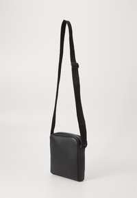 Bolso cruzado de cuero negro con forma rectangular, textura suave y correa negra ajustable. Presenta un cierre de cremallera en la parte superior.