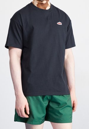 Osoba ubrana w czarny t-shirt z krótkim rękawem z małym czerwonym logo i zielone szorty, stojąca na jednolitym białym tle.
