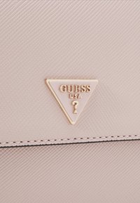 Světle růžová texturovaná kabelka s trojúhelníkovou zlatou plaketu s logem "GUESS USA" a akcentem otazníku, s tmavými švy.