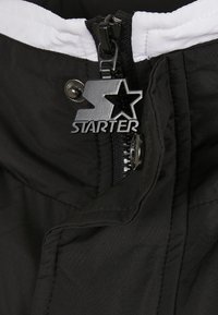 Chaqueta negra con tela texturizada, forro interior blanco y tirador de cremallera metálico con un logo de estrella.