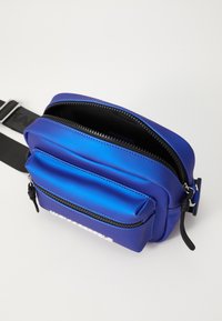 Blå crossbody-veske laget av glatt materiale, med en glidelåst hovedkompartiment og frontlomme, justerbar stropp, og hvit logo-detalj.