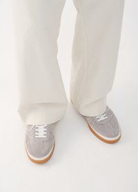 Grå suede sneakers med hvite snørebånd, hvite kanter og såler av gummigummi, kombinert med løstsittende lyse bukser.