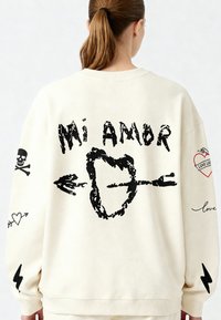 Felpa oversized color crema con testo grafico nero "Mi Amor", un design a cuore con freccia e motivi di teschi, cuori e fulmini.