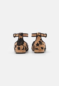 Pár béžových korkových plochých sandálů s černým květinovým vyšíváním a pásky kolem kotníků, zobrazené zezadu na bílém pozadí.