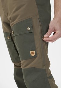 Vihreät ja ruskeat cargo-housut, joissa on taskut, vetoketjusulku ja metallinapit. Sisältää näkyvän merkkilapun jalassa. Tekstimäinen kangas.