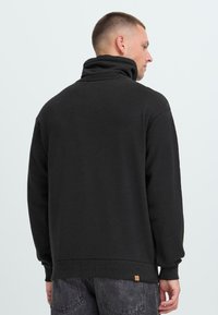 Hombre con cabello corto, vistiendo una sudadera negra de cuello alto y jeans de lavado oscuro, de espaldas contra un fondo claro y liso.