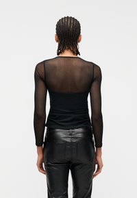 Άτομο με πλεξούδες που φοράει ένα διαφανές μαύρο μακρυμάνικο τοπ και μαύρο δερμάτινο παντελόνι, εμφανίζεται από πίσω σε απλό φόντο.