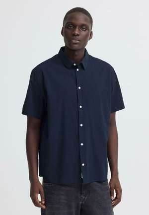 Joven con cabello corto que lleva una camisa azul marino de manga corta con botones y jeans gris oscuro, sobre un fondo claro y liso.