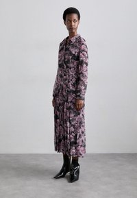Robe midi fleurie avec col, manches longues et jupe plissée en noir et rose. Portée avec des bottines noires à bout pointu. Fond minimaliste.