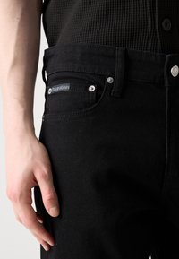 Crne traperice od denima sa srebrnim detaljima, s Calvin Klein oznakom na prednjoj manžetni i teksturiranim, prilagođenim dizajnom.