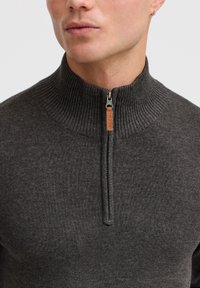 Dark grey zip-up sweater with a high ribbed collar, featuring a subtle texture and a small leather tag on the zip, showcasing a minimalist design.