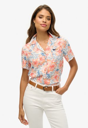 Chemise à manches courtes en coton blanc avec un motif floral rose, des accents bleus et un logo. Associée à un pantalon blanc et une ceinture marron.