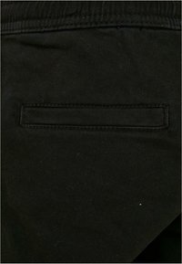 Nahaufnahme von schwarzen Stoffhosen mit elastischem Bund und einer horizontalen Paspeltasche auf der Rückseite.