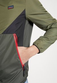 Zelená bunda s černými akcenty, vybavená kapsou na půl zip s červeným zipem. Vyrobena z lehkého, strukturovaného materiálu s elastickými manžetami.