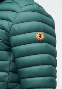 Μανίκι μπουφάν σε πράσινο τιρκουάζ με στρογγυλό πορτοκαλί ένθετο που απεικονίζει ένα παπί που τραγουδά και την επιγραφή "SAVE THE DUCK".