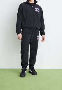 Svart träningsdräkt med en jacka med dragkedja och byxor med elastisk midja, båda med färgglada grafiska tryck. Bärs med svarta sneakers.