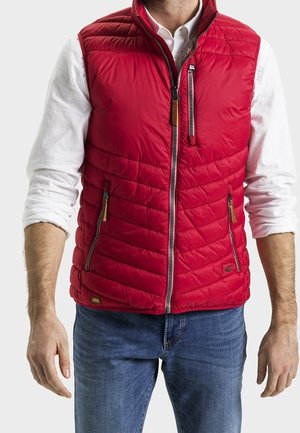 Mand iført en rød quiltet vest over en hvid langærmet skjorte og blå jeans, stående med armene afslappet langs siden.