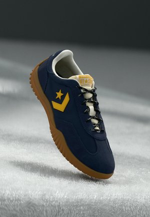 Tumed sinine tennis, mille suupper on sametist, kollased tähed ja triibud, valge keel ja tekstureeritud kummist tald. Mustad paelad on ühtlaselt läbi keeratud.