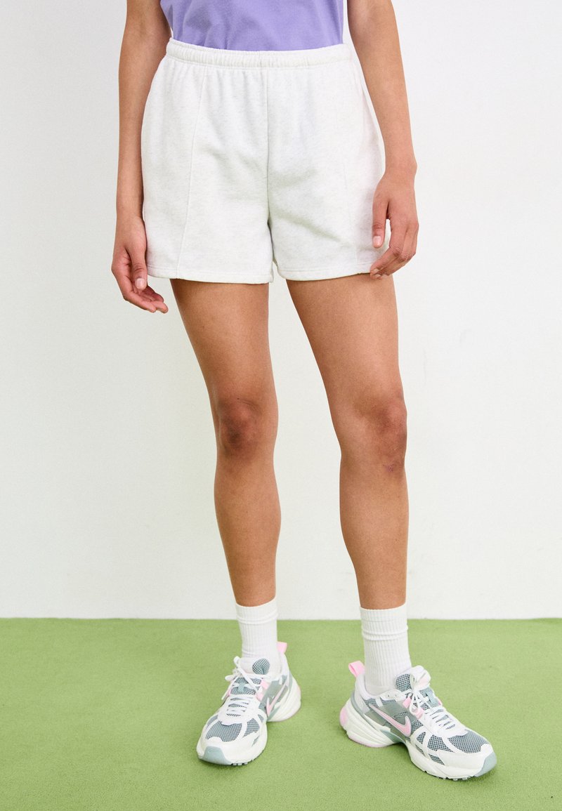 Shorts en coton gris clair avec une taille élastique, associés à des chaussures de sport grises et roses et des chaussettes blanches mi-mollet. Sol en texture verte.