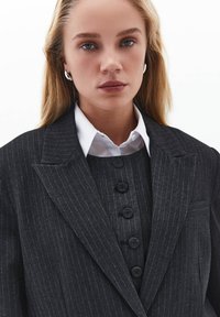 Chaqueta de traje a rayas oscuras con solapas con muesca, llevada sobre una camisa blanca con cuello. Presenta un chaleco gris ajustado con cinco botones redondos.