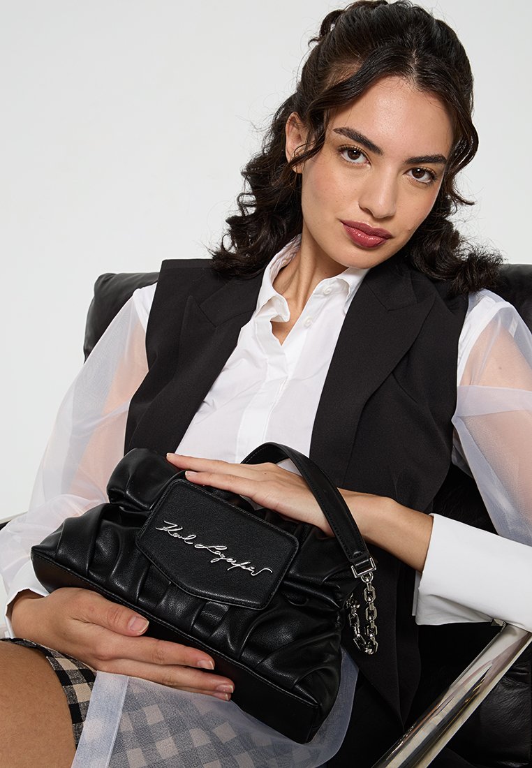 Bolso de mano negro, acolchado, hecho de cuero sintético, con un parche de logo en la parte frontal y una correa acentuada con cadenas, sostenido por una mujer con una camisa blanca y un chaleco.