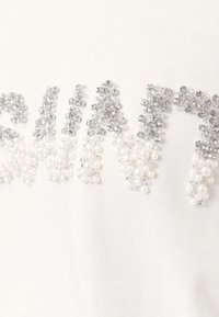 Biele tričko s vystúpenými písmenami zdobenými priehľadnými a perlovými korálikmi, ktoré vytvárajú textúrovaný povrch s trblietavým efektom.