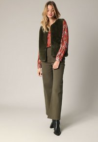 Olivgrüne Kunstfellweste über einer gemusterten roten Bluse, kombiniert mit olivgrünen Hosen und schwarzen Ankle Boots, Ganzkörperaufnahme.