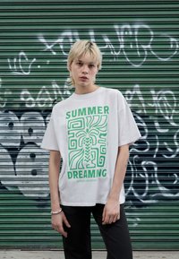 Λευκό βαμβακερό t-shirt με πράσινο γραφικό σχέδιο που αναγράφει "SUMMER DREAMING," αφηρημένα σχήματα και έναν φοίνικα. Χαλαρή εφαρμογή με κοντά μανίκια.