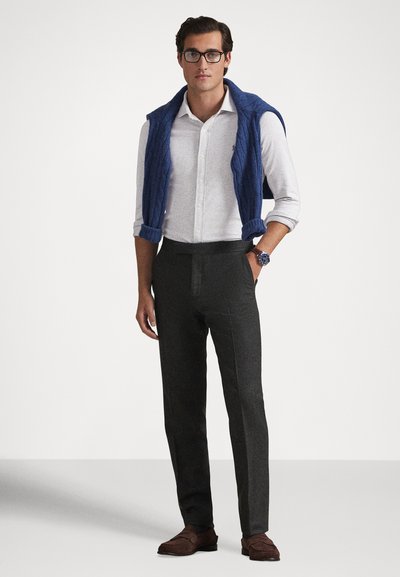 Άνδρας με γυαλιά, λευκό πουκάμισο με κουμπιά και γυρισμένα μανίκια, μπλε πουλόβερ περασμένο στους ώμους, σκούρο παντελόνι, καφέ παπούτσια τύπου loafer και ρολόι χειρός.