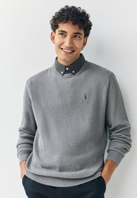 Grå ribbstickad tröja med rund halsringning, bärs över en svartvit rutig skjorta med krage, med en liten logotyp på bröstet.