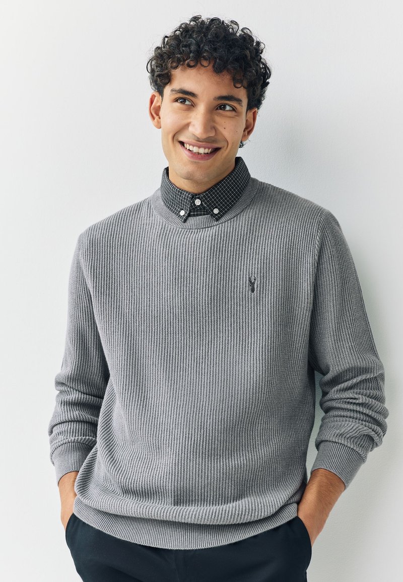 Grå ribbstickad tröja med rund halsringning, bärs över en svartvit rutig skjorta med krage, med en liten logotyp på bröstet.