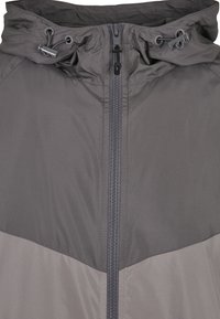 Grå zip-up jakke med en kontrasterende mørkere grå panel, justerbar hætte med snørremateriale og en glat, mat stoftekstur.