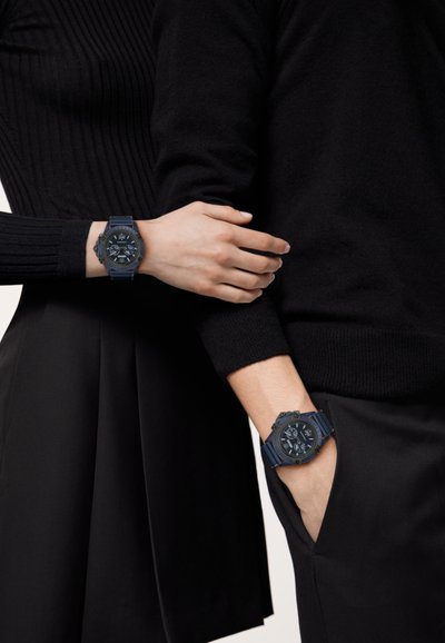 Deux montres bleues avec des bracelets en caoutchouc texturé et des cadrans sombres sont portées aux poignets de personnes vêtues de noir.
