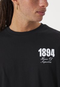 Tricou negru cu un grafic alb care include textul "1894" și "House Of Superbia". Materialul este moale, având un decolteu clasic tip crew.