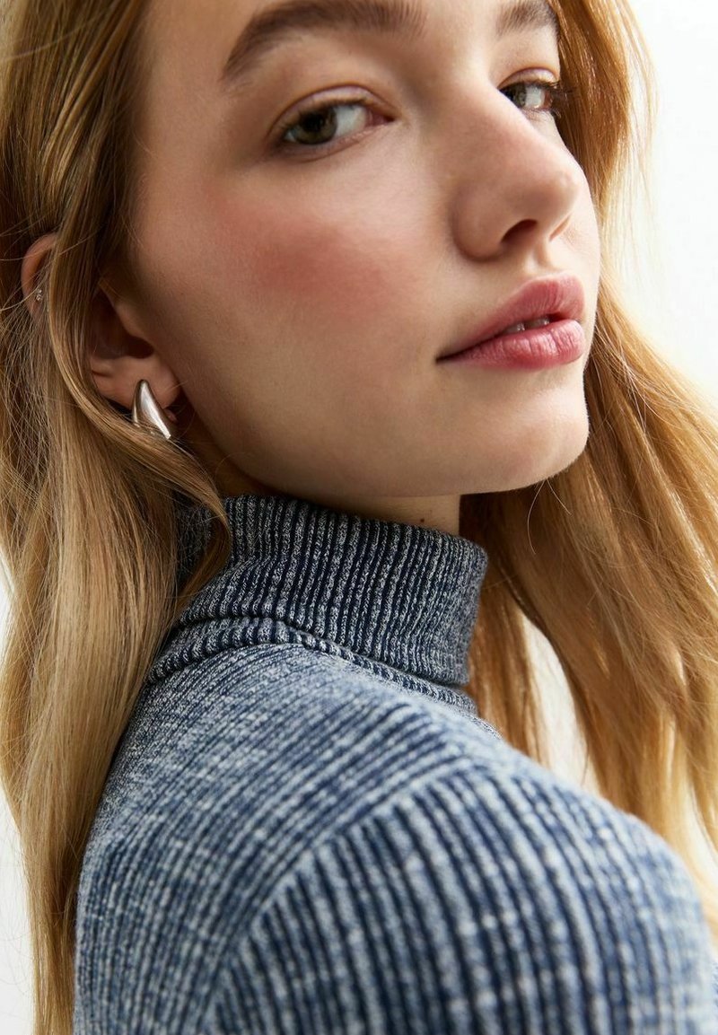 Pullover blu con collo a lupetto e righe verticali. Modello con orecchino argento; capelli lisci cadono sulla spalla.