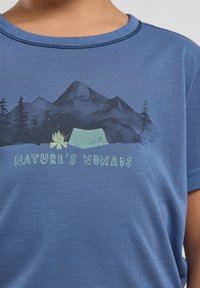 T-shirt azul de manga curta com um gráfico de montanhas, uma tenda e uma fogueira, com o texto "OS NÔMADES DA NATUREZA" em verde.