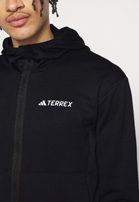 Felpa nera Terrex con zip, colletto alto e logo ricamato. Texture liscia con un design elegante e aderente.