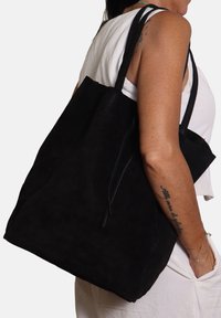 Donna che indossa una maglietta senza maniche bianca e pantaloni, con una grande borsa a tracolla in suede nera, mostrando un tatuaggio sul braccio e un bracciale.