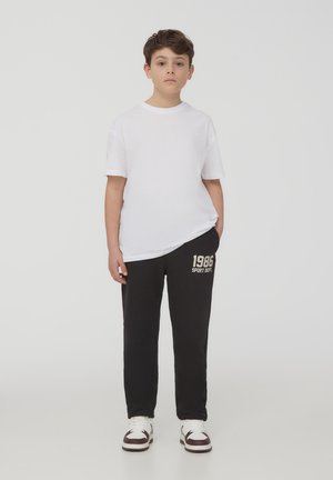 Biała bawełniana koszulka połączona z czarnymi dresami z złotym napisem '1986 SPORT DEPT.', dopełniona białymi i burgundowymi trampkami.