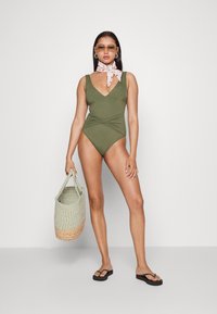 Fato de banho verde-oliva de uma só peça, com um decote em V profundo, detalhes franzidos e pernas altas. Combinado com um lenço estampado e uma bolsa tecida.
