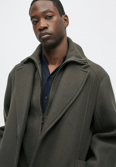 Olīvu zaļš mētelis ar augstu apkakli un rāvējslēdzēju priekšā, uzlikts virs tumši zila krekla. Mētelis ir ar teksturētu audumu un strukturētu dizainu.