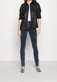 Svart läder skjortjacka med två bröstfickor, bärs över en vit topp, matchad med mörkblå slim-fit jeans och svarta flip-flops.