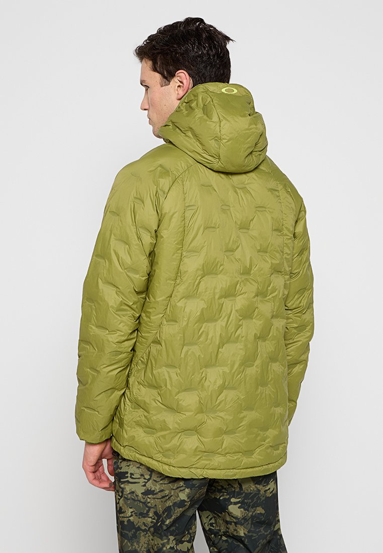 Chaqueta verde aislante con un diseño acolchado y capucha. Presenta una textura suave y un detalle de logo en la parte trasera. Se lleva con pantalones de camuflaje.