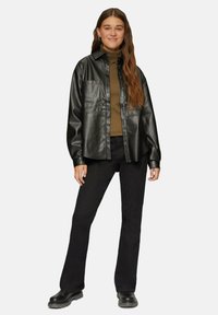 Schwarze Kunstlederhemdjacke mit Druckknöpfen, zwei Vorderaschen, getragen über einem braunen Rollkragenpullover, kombiniert mit schwarzen Bootcut-Hosen und Stiefeln.