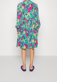 Abito floreale dai colori vivaci, con una base verde e motivi rosa e gialli. Maniche lunghe, vita sgambata e lunghezza al ginocchio.